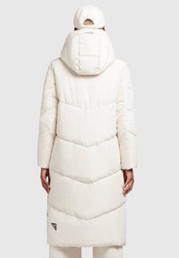 Manteau doudoune blanc long avec capuche. Présente un matelassage en chevrons et un écusson logo en bas à droite. Texture douce et conception longueur genoux.