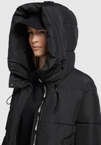 Schwarze Puffjacke mit einer großen Kapuze, ausgestattet mit Kordelzügen, einem durchgehenden Reißverschluss und einem Logo-Design entlang der inneren Reißverschlusslinie.