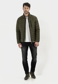 Olivenfarbene Pufferjacke mit horizontalem Steppmuster, Reißverschluss vorne und hohem Kragen, getragen über einem weißen Hemd und schwarzen Jeans.