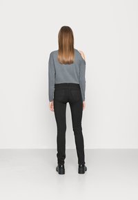 Mulher com cabelo castanho liso, vestindo uma blusa cinza de ombros à mostra, calças justas pretas e sapatos plataforma pretos, de pé, virada para uma parede branca.