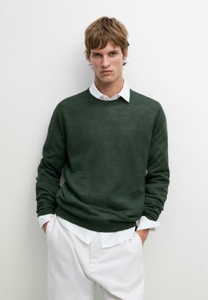 Giovane uomo con maglione verde scuro sopra una camicia bianca con colletto e pantaloni bianchi, in piedi contro un muro chiaro e semplice con le mani nelle tasche.