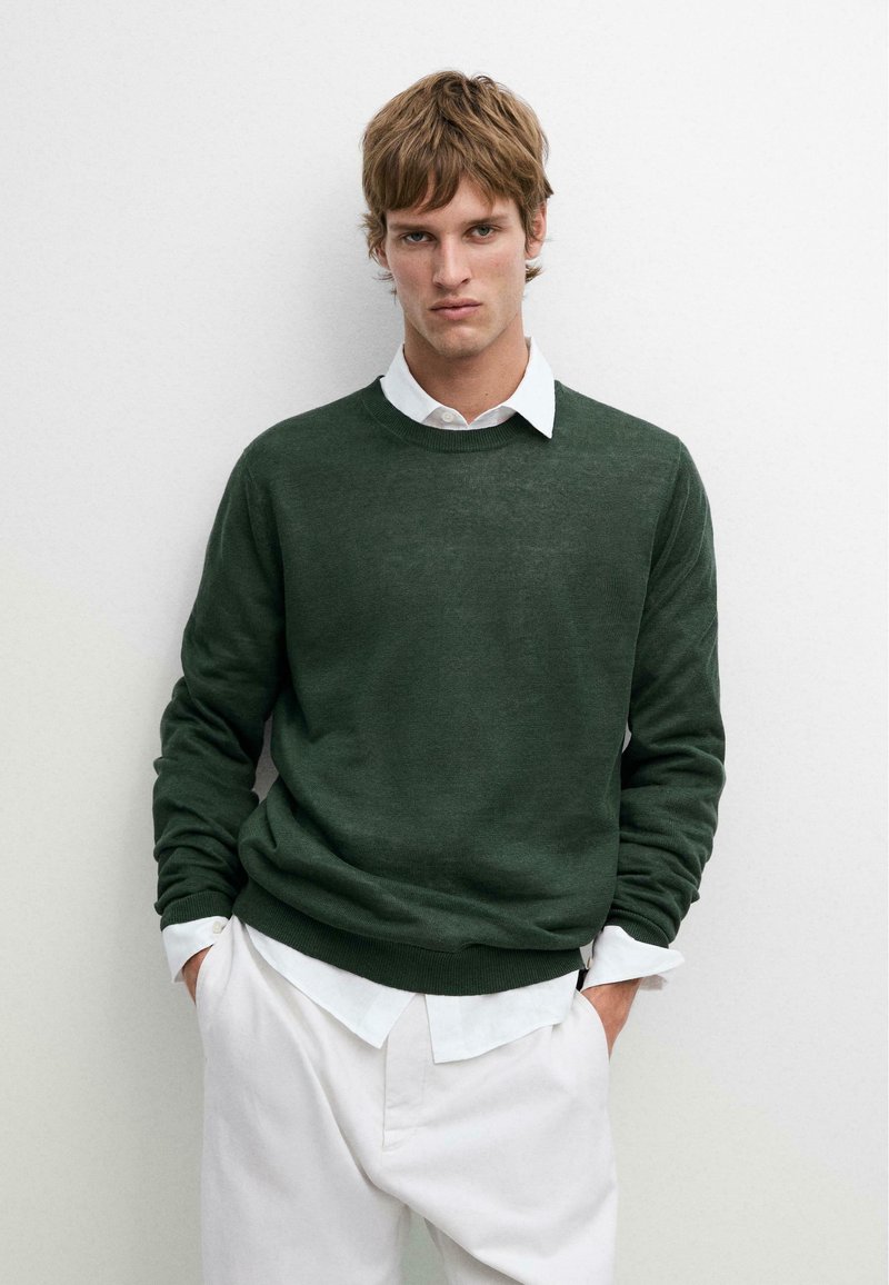 Junger Mann trägt einen dunkelgrünen Pullover über einem weißen Hemd mit Kragen und weiße Hose, steht vor einer schlichten hellen Wand mit den Händen in den Taschen.