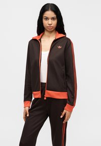 Καφέ αθλητικό μπουφάν με πορτοκαλί φερμουάρ και γιακά, που διαθέτει τρεις πορτοκαλί ρίγες στα μανίκια και ένα μικρό λογότυπο στο στήθος.