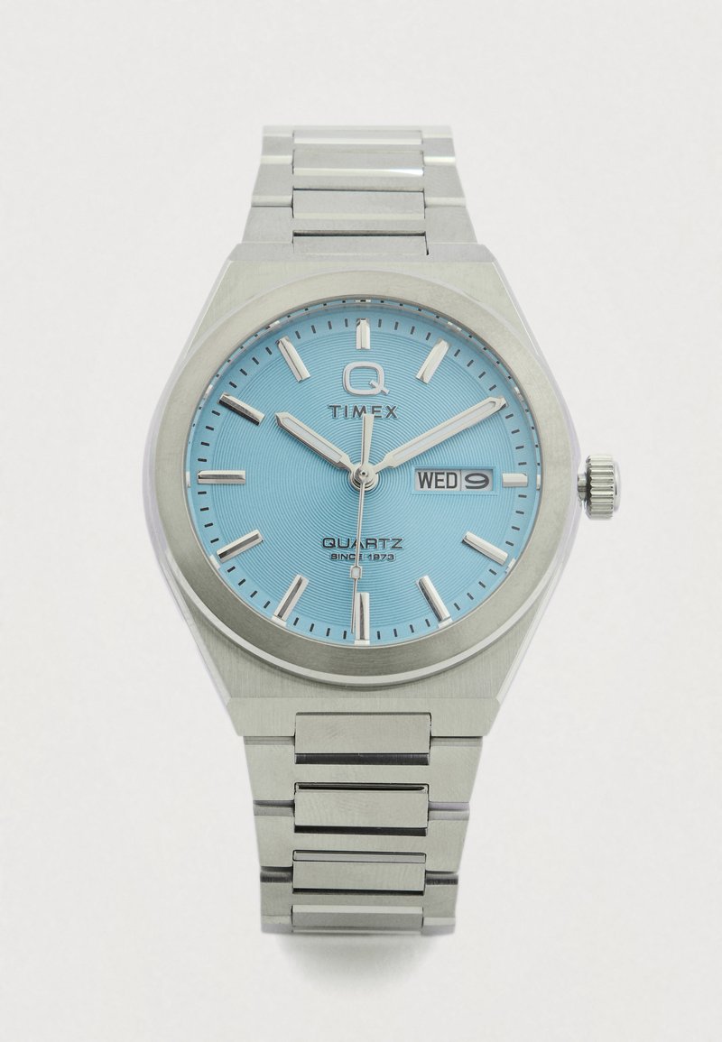 Kvarcinis laikrodis su šviesiai mėlynu ciferblatu, sidabriniais valandų žymekliais ir dienos/data rodmenimis. Nerūdijančio plieno korpusas ir apyrankė su šlifuotu paviršiumi.