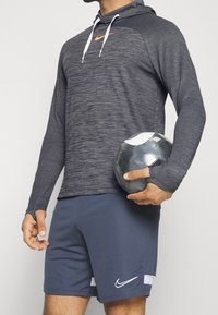 Dunkelgrauer sportlicher Kapuzenpullover mit orangefarbener Nike-Logo, langen Ärmeln und Daumenlöchern, kombiniert mit navyblauen Shorts mit weißen Nike-Akzenten. Hält einen Fußball.