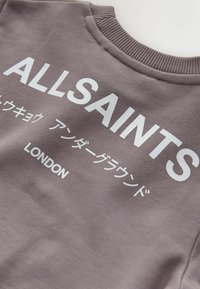 Pelēks džemperis ar baltu tekstu "ALLSAINTS," papildināts ar japāņu rakstzīmēm un "LONDON" apakšā. Ribu apkakles detaļa.