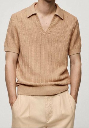 Maglione polo beige a maniche corte con scollatura a V, caratterizzato da un motivo a maglia texture e orli e polsini a costine. Indossato sopra pantaloni chiari.