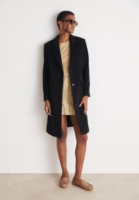 Manteau en laine noir avec revers, top beige, jupe métallique dorée et ballerines brunes à motif ; accessoirisé avec des lunettes de soleil et des boucles d'oreilles.