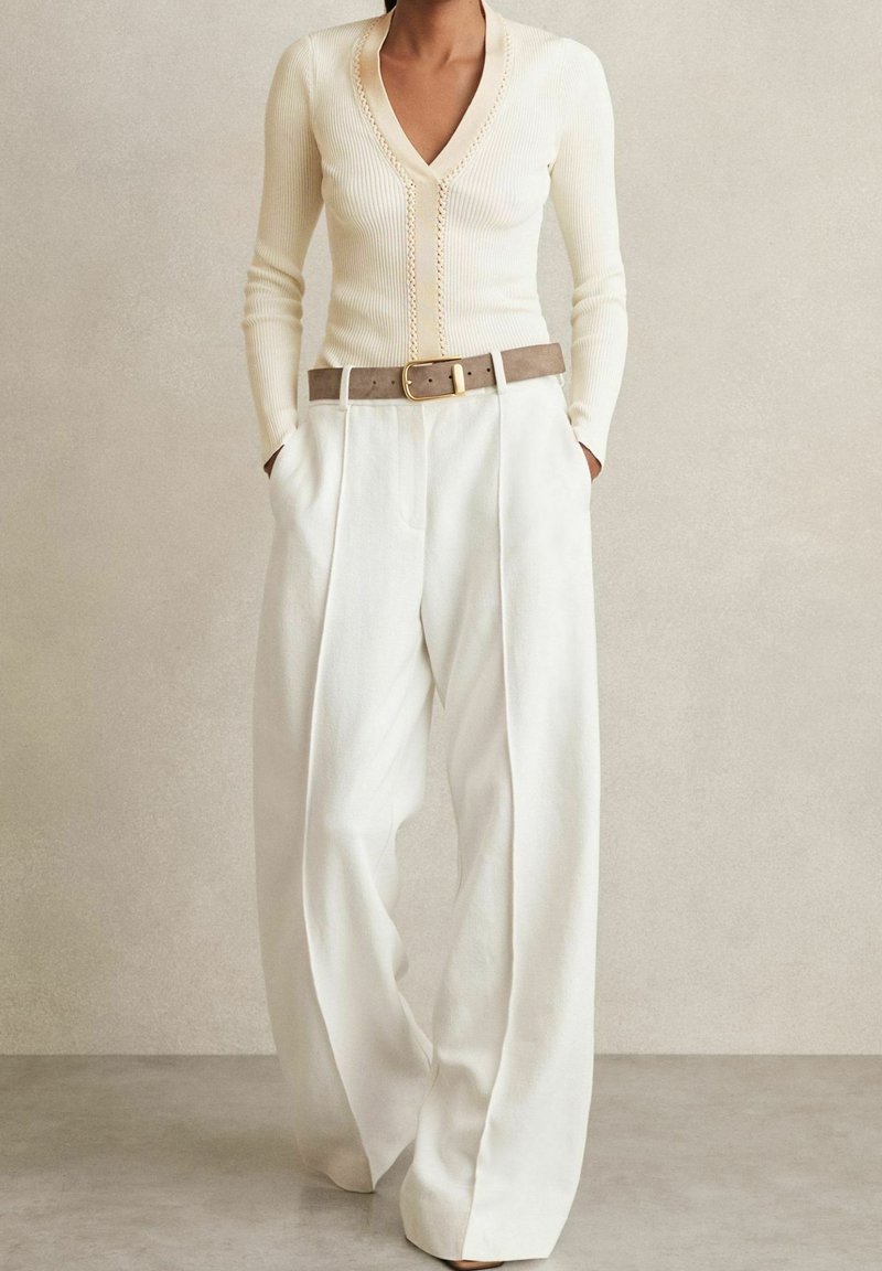 Personne portant un pull côtelé crème à col en V rentré dans un pantalon blanc taille haute à jambes larges avec une ceinture marron clair et les mains dans les poches.