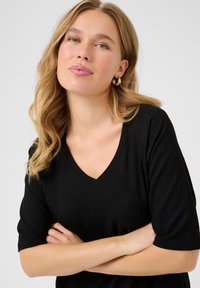 Top nero con scollo a V, maniche corte, in tessuto a coste e vestibilità rilassata. Il modello posa con le braccia incrociate, evidenziando il design dello scollo e delle maniche.