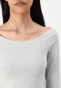 Svijetlosiva, prilagodljiva majica dugih rukava s širokim izrezom off-the-shoulder; napravljena od mekog, teksturiranog materijala s suptilnim šavnim detaljima.
