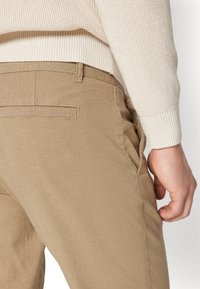 Pantaloni in cotone beige con una texture liscia, dotati di una tasca posteriore e un fit slim, abbinati a un maglione lavorato a maglia color crema.