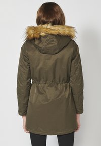 Olivovo zelená parka s sťahovacím pásom, ktorá má odnímateľnú hnedú kožušinovú kapucňu a hladkú textúru, navrhnutá pre teplo.