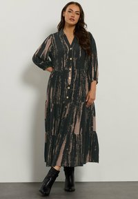 Langes, knöpfbares Kleid in dunklem Grün mit abstrakten pinken und cremefarbenen Mustern. Hat einen ausgestellten Rock und Dreiviertelärmel. Wird mit schwarzen Ankle Boots getragen.