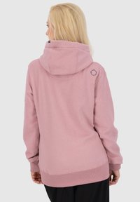 Donna con capelli biondi che indossa una felpa di un rosa polveroso con un logo testuale circolare vicino alla spalla destra, rivolta allontanandosi dalla macchina fotografica.