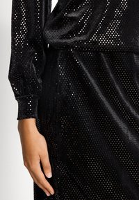 Svart sammetklänning med ett flertal små reflekterande prickar, långa ärmar och rynkade manschetter, vilket betonar struktur och glans.