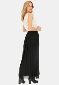 Czarna maxi spódnica z plisowanego materiału opada na ziemię. Połączona z białym bezrękawnikiem, posiadającym miękką teksturę i subtelny połysk.