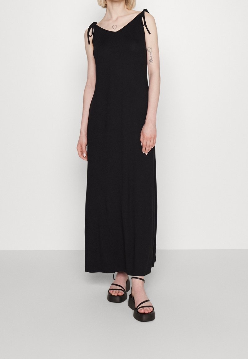 Femme portant une longue robe noire sans manches avec des bretelles à nouer et des sandales compensées noires, debout devant un fond clair uni.