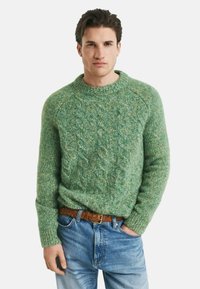 Homme portant un pull vert en maille torsadée, un jean bleu et une ceinture tressée marron, se tenant avec une main dans la poche devant un fond blanc.