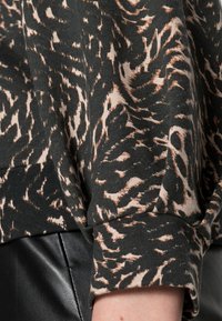 Tela con patrón en negro y marrón, que presenta una textura suave, con un diseño acogedor y un detalle de puño visible. Combinada con una falda de cuero negra.
