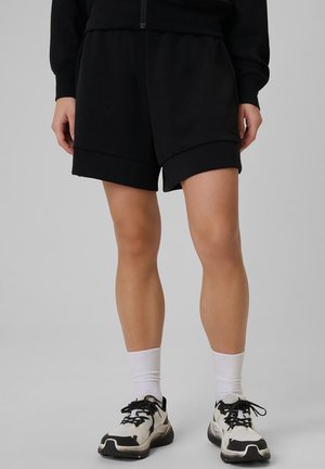 Persona con pantalones cortos negros, calcetines blancos hasta el tobillo y zapatillas deportivas negras y blancas, de pie frente a un fondo gris liso.