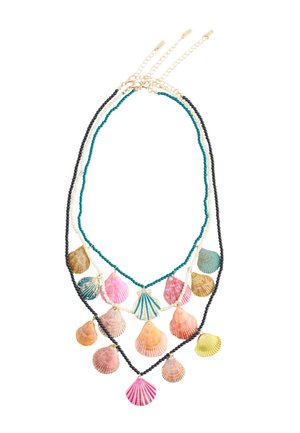 Frau trägt ein Bikinioberteil mit Strandmotiv und geschichtete bunte Muschelketten sowie passende Muschelohrringe vor einer hellen Wand.