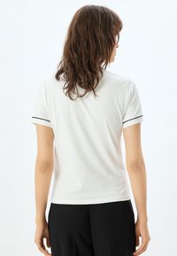 Camisa polo de manga corta blanca con acentos de rayas negras en las mangas, con un corte clásico y una textura de algodón suave, vista desde atrás.