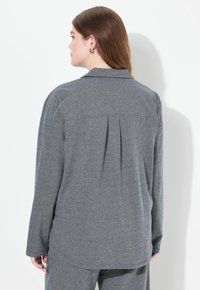 Camisa gris de manga larga con un patrón de espiga. Cuenta con un pliegue en la parte posterior, un cuello con solapa y un dobladillo redondeado. Está hecha de una tela texturizada.