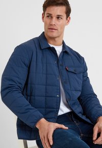 Navy blue quilted jacket featuring a button-up front, a chest pocket with flap, and a classic collar. Made from lightweight material.