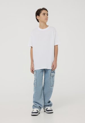 T-shirt bianco in cotone a maniche corte, abbinato a jeans cargo larghi azzurri chiari con tasche. L'outfit include sneakers bianche e nere.