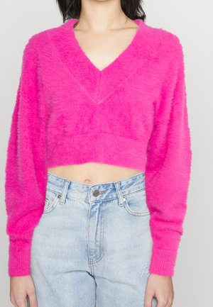 Maglione corto fuzzy di un brillante colore rosa, con scollo a V e maniche ampie, abbinato a jeans chiari a vita alta. Tessuto morbido e vestibilità ampia.