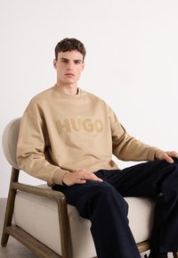 Sweatshirt beige avec le texte "HUGO" dans une teinte assortie, col rond et manches longues. Le modèle est assis sur une chaise avec un pantalon foncé.