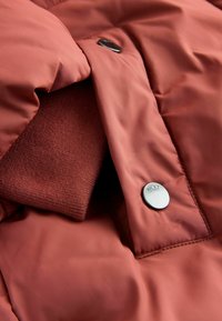 Rostfarbene Puffjacke mit glatter, glänzender Außenseite. Verfügt über ein strukturiertes Innenfutter und einen metallischen Druckknopf mit der Aufschrift "NEXT".