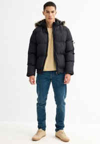 Chaqueta de plumas negra con capucha de pelo, que incluye acolchado para aislamiento, combinada con una camiseta beige y jeans azules. El calzado casual es beige.