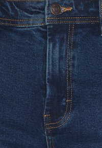 Temno modri jeans iz denima z sprednjo žepom z zlatim šivom, z metalnim gumbom za zapiranje in gladko teksturo z rahlo sijajem.