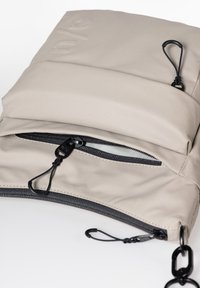 Beige strukturierte Tasche mit glatter Oberfläche, ausgestattet mit einem schwarzen Reißverschluss und einer Kettenriemen. Enthält eine Vorderseite mit Reißverschlusstasche, die mit Ziehschlaufen versehen ist.