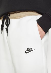 Hånd i lommen på hvide Nike joggingbukser med sort snor, beige taljebånd og sort langærmet top synlig ved taljen.