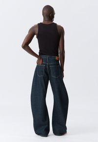 Tamnoplave izrezane traperice širokih nogavica s visokim strukom, stražnjim džepom i vidljivim detaljima šava, uparene s crnim majicama bez rukava.