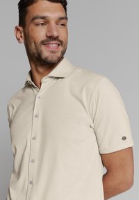 Beige polo shirt met korte mouwen, gemaakt van zachte stof, met een klassieke kraag, knoopsluiting en een klein logo-accent op de mouw.