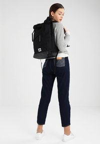 Mochila negra tipo roll-top con correas ajustables, parche de logo blanco y bolsillo con cremallera lateral. Lleva puesta con pantalones de mezclilla y zapatillas blancas.