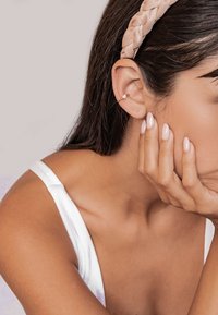 Woman with braided headband resting her face on her hand, showing gold ear cuff and light pink polished nails.