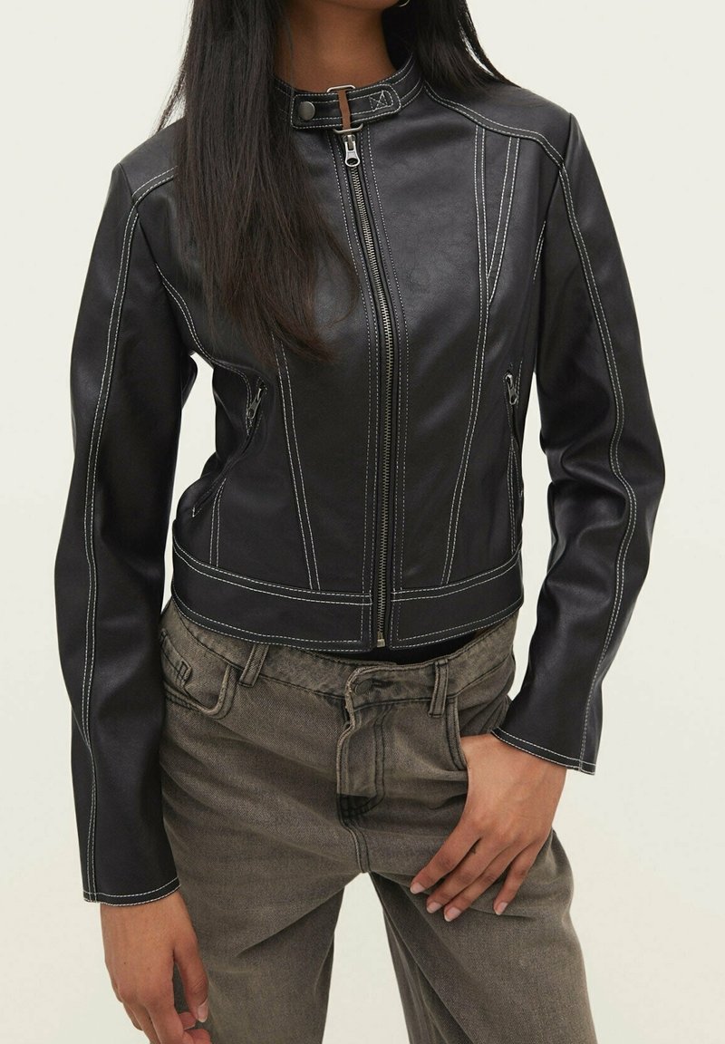 Black leather jacket with white stitching, high collar, front zip, and angled side pockets. Paired with beige high-waisted pants.