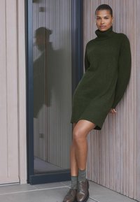 Dunkelgrünes, geripptes Rollkragenkleid mit lockerem Schnitt, kombiniert mit gestreiften Socken und braunen Schnürschuhen vor einer holzvertäfelten Wand.