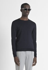 Pullover navy in maglia con collo tondo e maniche lunghe, abbinato a pantaloni grigi chiari. Il modello indossa occhiali da sole neri a forma quadrata.