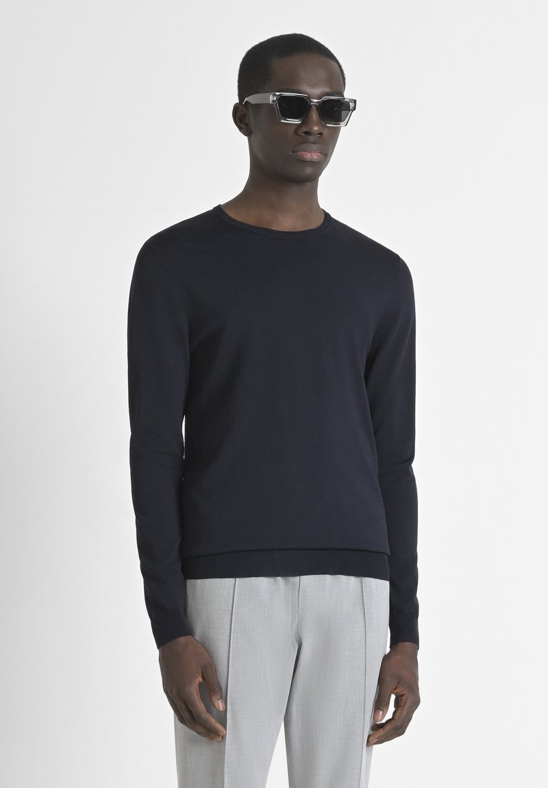 Pullover navy in maglia con collo tondo e maniche lunghe, abbinato a pantaloni grigi chiari. Il modello indossa occhiali da sole neri a forma quadrata.