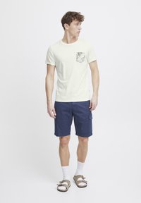 Una camiseta de manga corta de color claro con un bolsillo que presenta un patrón tropical, combinada con pantalones cortos cargo azules y sandalias marrones.