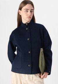 Marinefarbener Wollmantel mit hohem Kragen, vorne mit Knopfverschluss und zwei vorderen Taschen. Kombiniert mit einer olivgrünen Clutch und einem Paillettenrock.