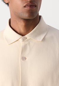 Polo beige con una consistenza morbida, caratterizzato da un colletto a punta e due bottoni al collo. Tessuto liscio con un leggero stretch.