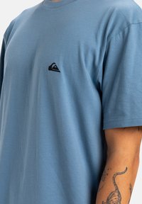 Camiseta de algodón azul claro con mangas cortas, escote redondo y un pequeño logotipo negro en el pecho. Textura suave, diseño casual.