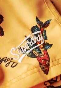 Tessuto giallo con il logo ricamato "Superdry", decorato con una stampa colorata di pesci e fiori. Dettagli di cucitura visibili lungo il bordo.